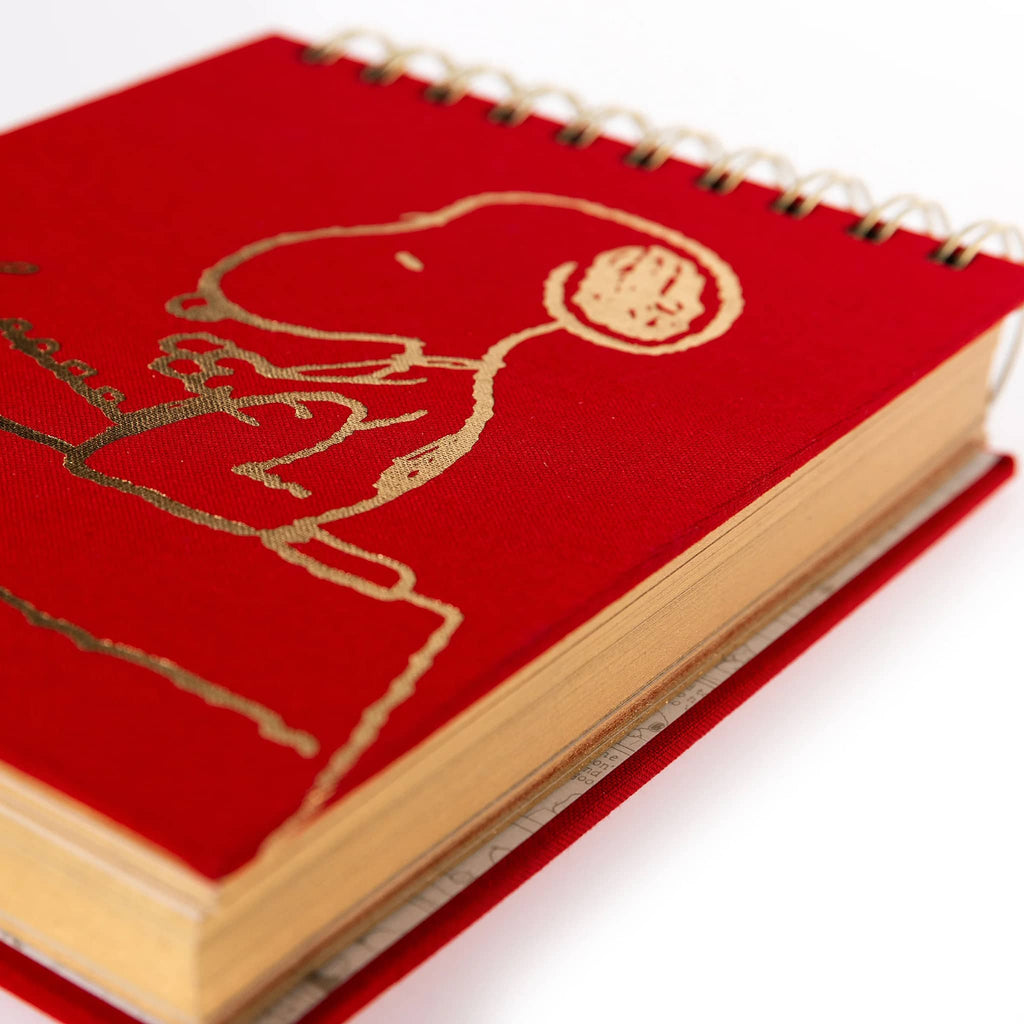 Red notebook with an illustration of Snoopy sitting on a desk with a typewriter in gold foil, detail of page edges with gold, on a white background.