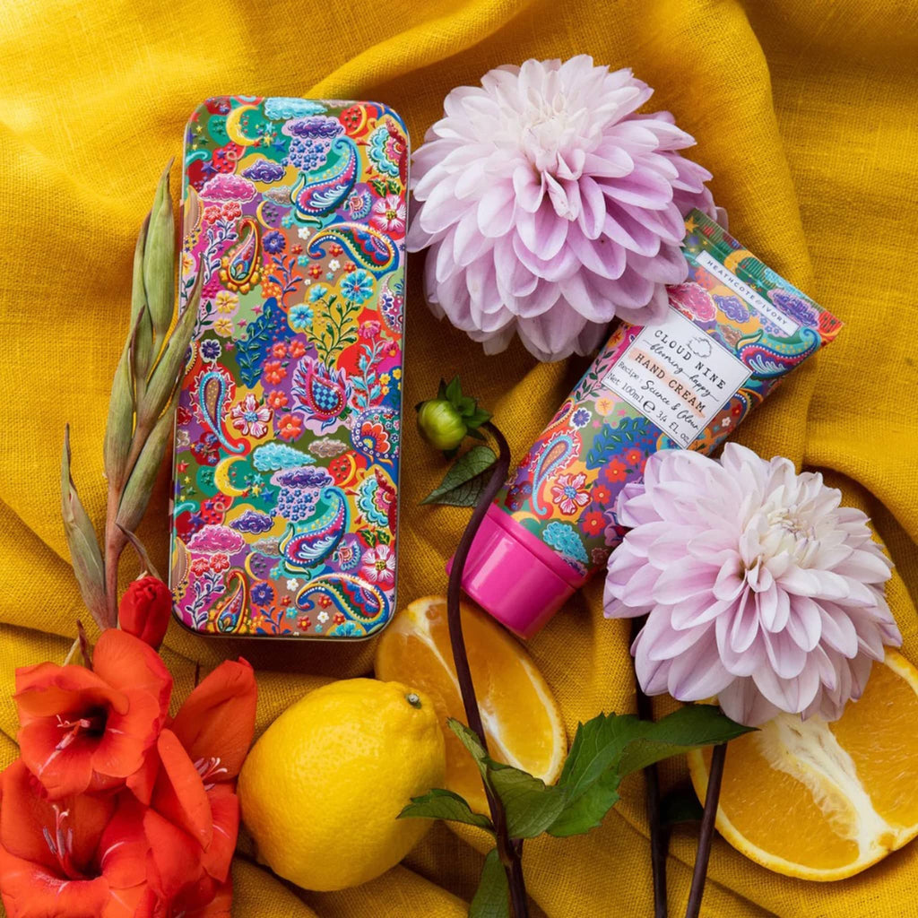 Heathcote & Ivory Cloud Nine Blooming Happy 3.4 ounce Hand Cream tube with collectible tin packaging, both with a colorful paisley print on a yellow fabric background with peonies and lemons.