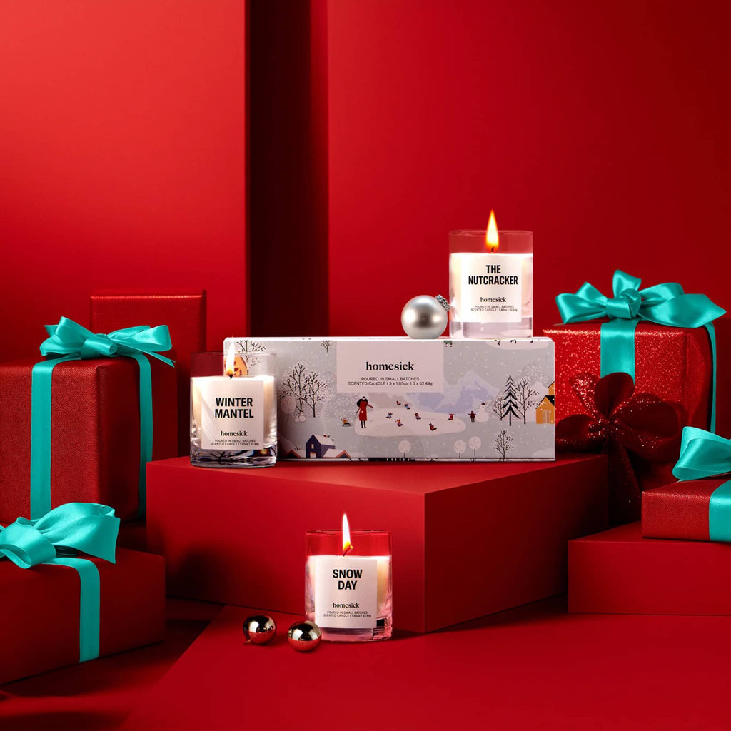 Decorative setup with three mini candles with box packaging and wrapped gifts on a red background.