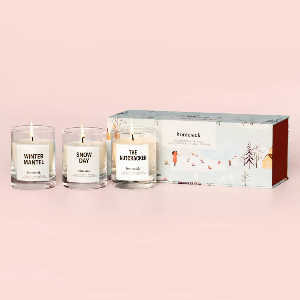 Three lit mini candles with labels and winter illustrated box packaging on a pink background.