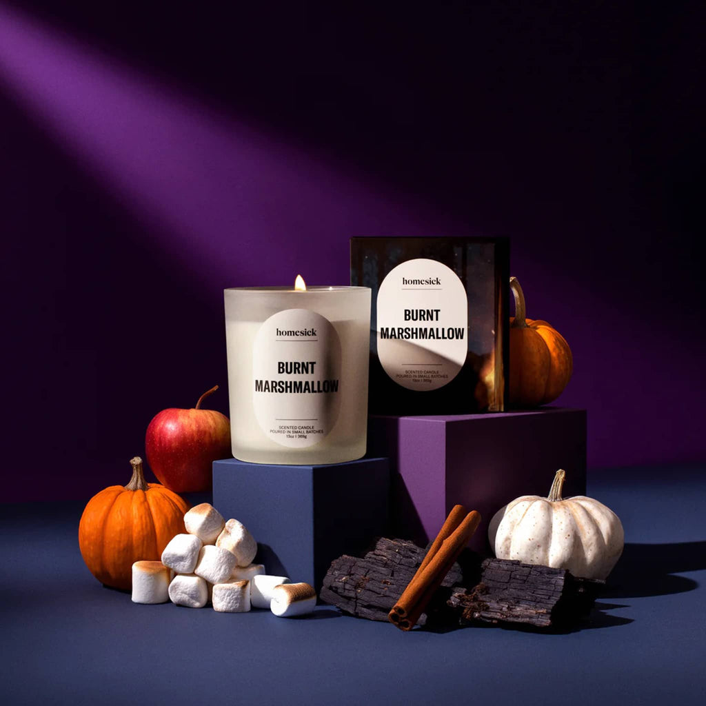 Homesick 'Burnt Marshmallow' candle with packaging, pumpkins, and marshmallows on a purple background.