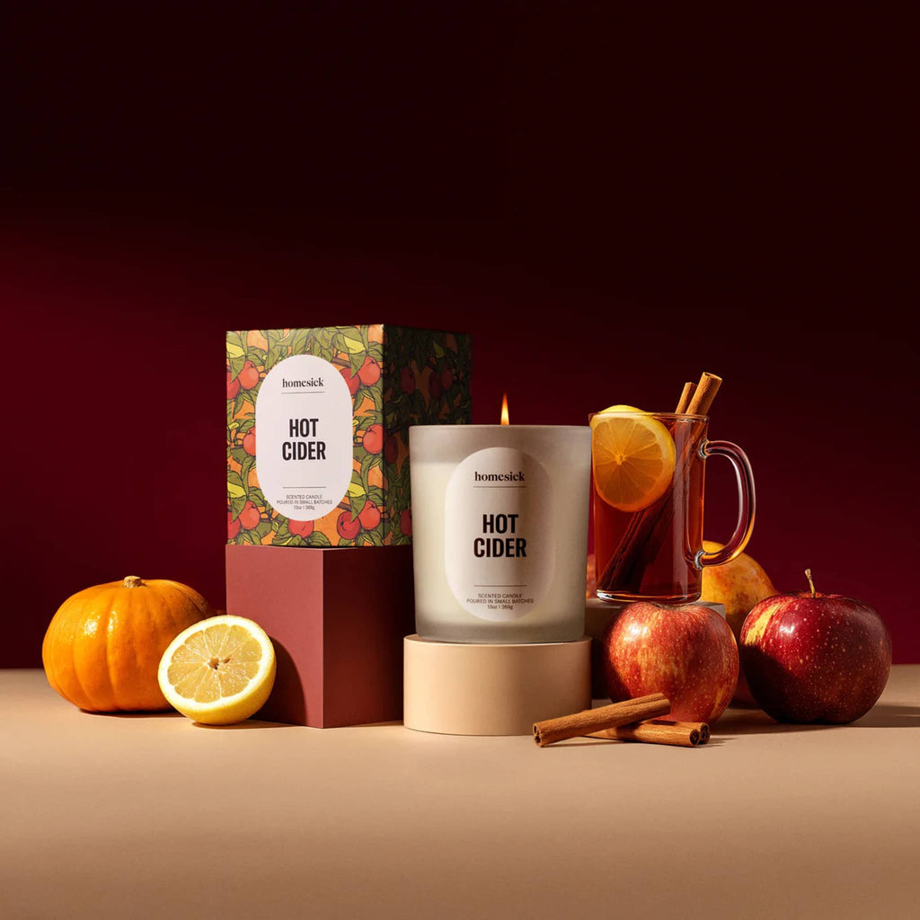 Homesick Hot Cider candle with packaging, apples, oranges, and cinnamon sticks on a warm background.