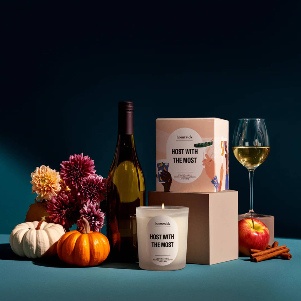 Homesick Host with the Most candle and box with wine bottle, pumpkins, flowers, apple, cinnamon sticks and a glass of white wine on a blue surface with a dark background.
