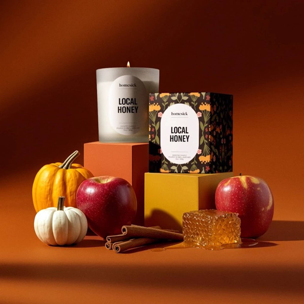 Homesick Local Honey candle with box and apples, pumpkins, cinnamon sticks and honeycomb on an orange background.