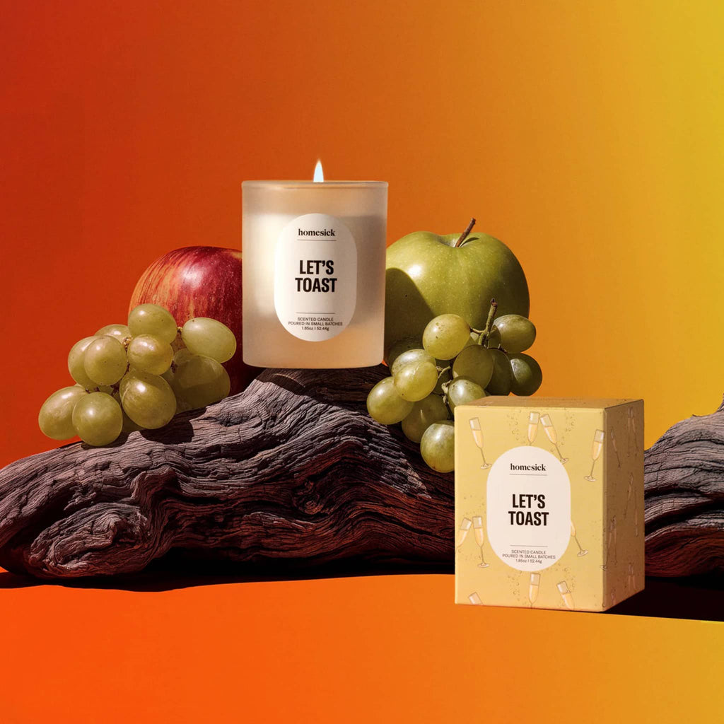 Mini candle labeled 'Let's Toast' with fruits and a box on an orange ombre background.