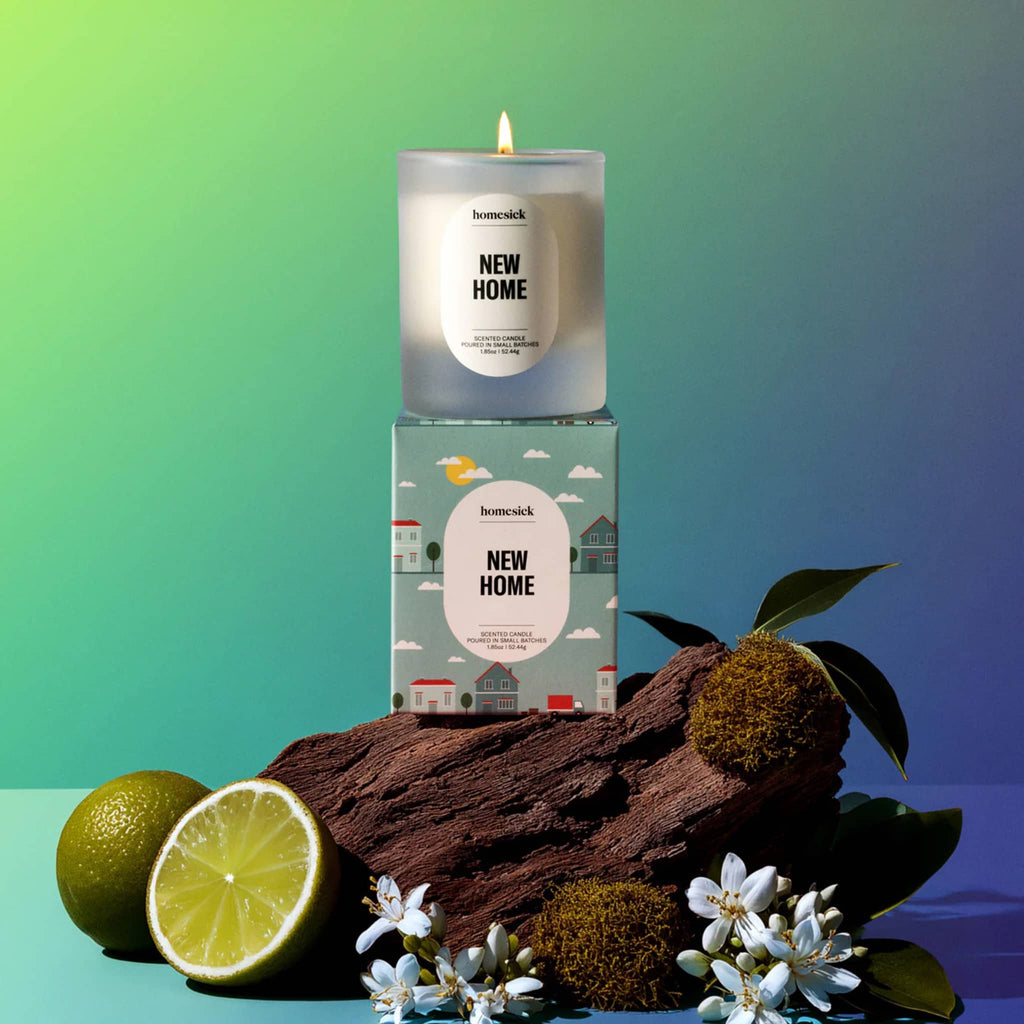 Homesick 'New Home' mini candle and packaging on a wooden block with limes and flowers against a gradient background.