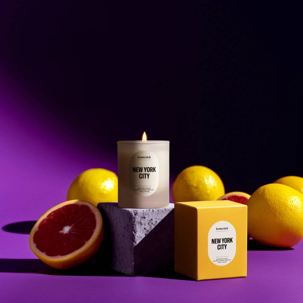 Homesick New York City mini scented candle on cement block, wick lit, with yellow box and lemons on a purple background.