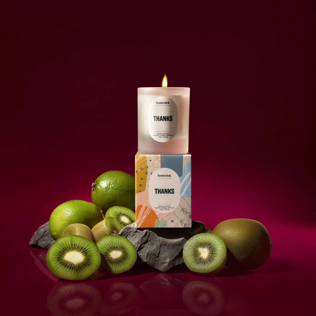 Mini candle with 'THANKS' and box packaging surrounded by kiwis, limes and wood on a red background.