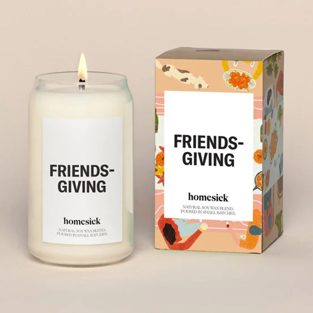 Homesick Friendsgiving scented soy wax blend candle in can-shaped glass with lit wick, next to illustrated orange gift box packaging.