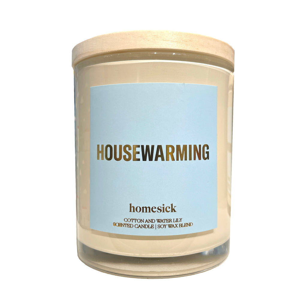 Homesick Housewarming Starter Collection candle in a clear glass tumbler with wood lid and a light blue label with "housewarming" in gold foil lettering along with the logo.
