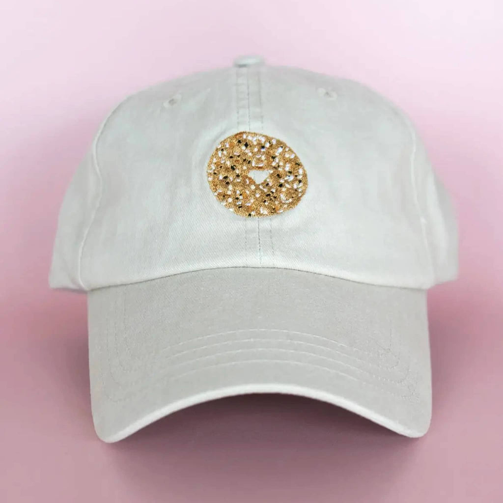 Light khaki baseball cap with an everything bagel embroidered on the front, on a pink background.