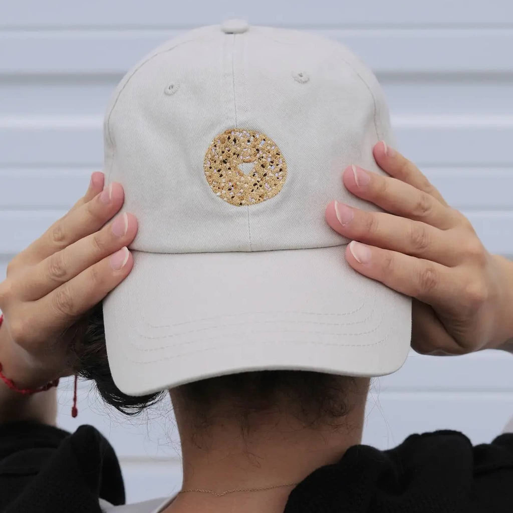 Light khaki baseball cap with an everything bagel embroidered on the front, shown backwards on a person with hands holding it.
