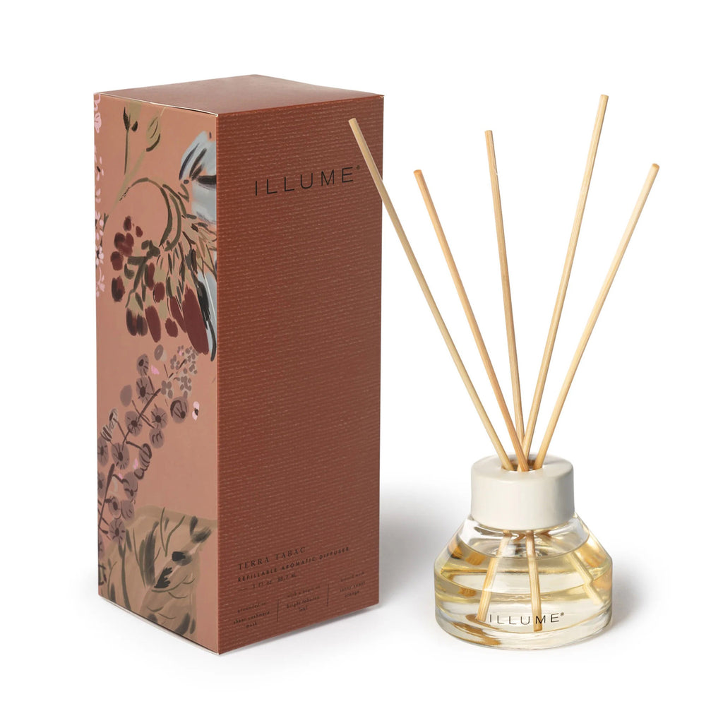 Illume Terra Tabac refillable aromatic reed diffuser in modern glass jar with white ceramic top beside rust colored gift box with floral illustrations.