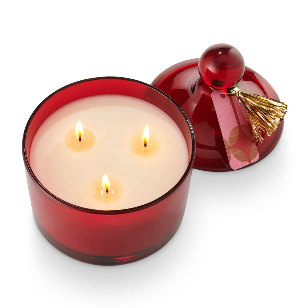 Illume Memory Lane Mistletoe Kisses scented soy wax holiday candle in dark red lidded glass vessel with gold tassel, overhead view with 3 wicks lit and lid rested beside.