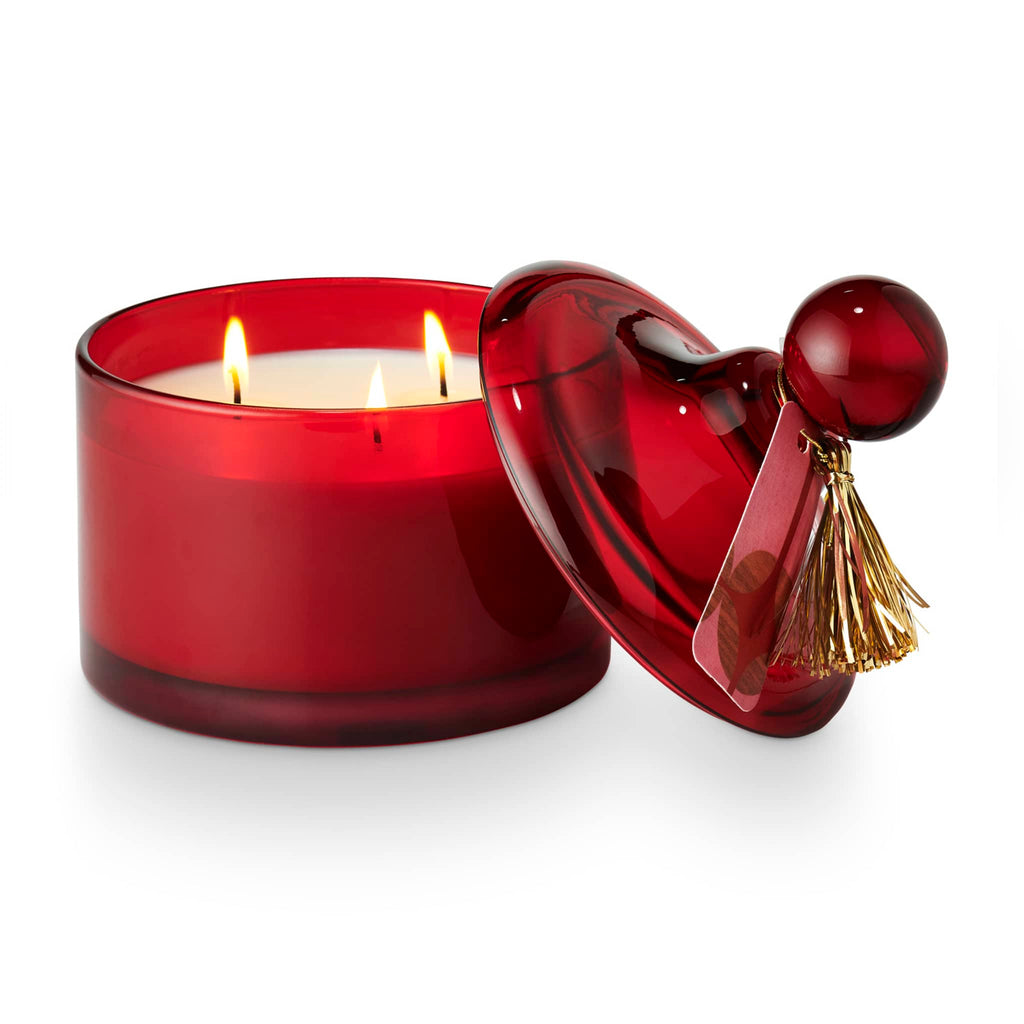 Illume Memory Lane Mistletoe Kisses scented soy wax holiday candle in dark red lidded glass vessel with 3 wicks lit and lid resting against the side.