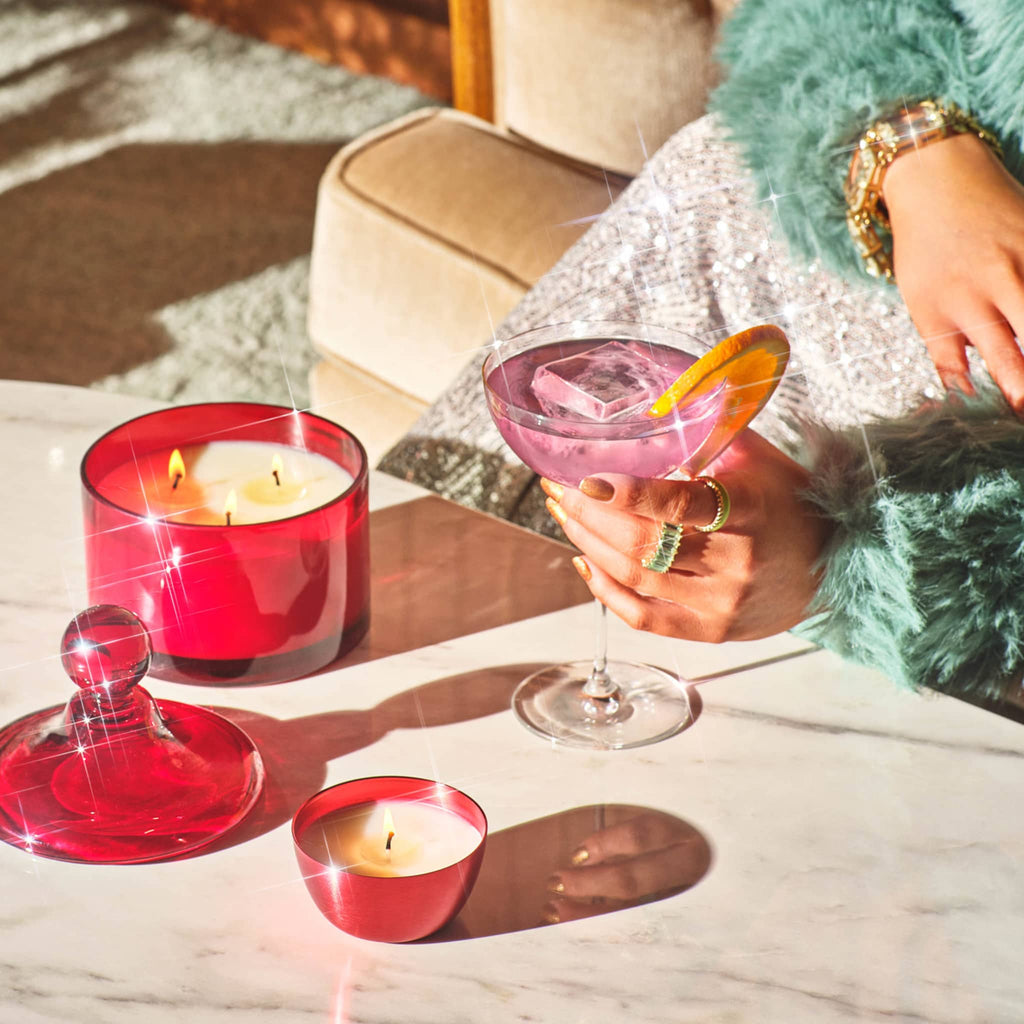 Illume Memory Lane Mistletoe Kisses scented soy wax holiday candle in dark red lidded glass vessel with gold tassel beside the red tin version and a martini glass.