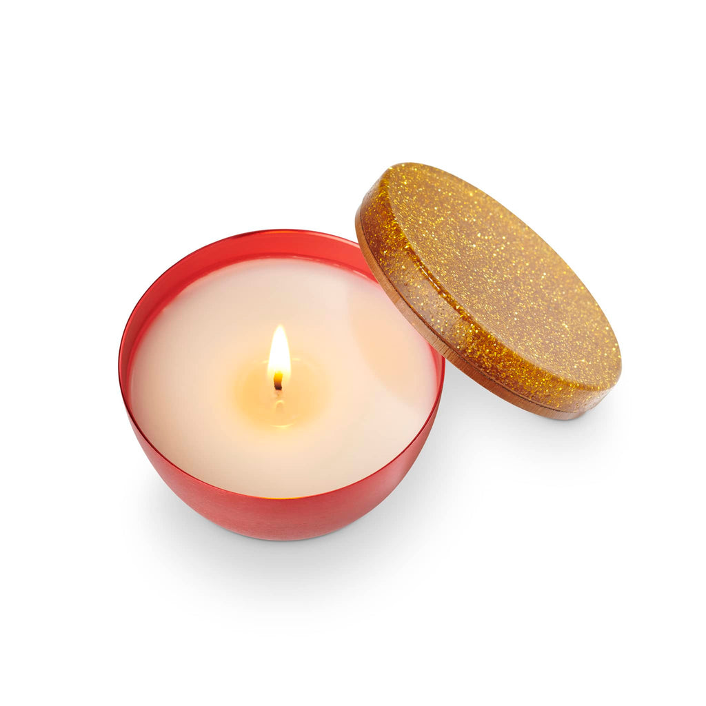 Illume Memory Lane Mistletoe Kisses holiday scented soy wax candle in red metal vessel with glittery gold lid, lid off and wick is lit.