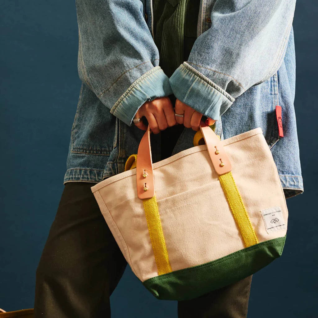 Person holding a natural canvas tote bag with green base, chartreuse straps and leather handles against a dark blue background.