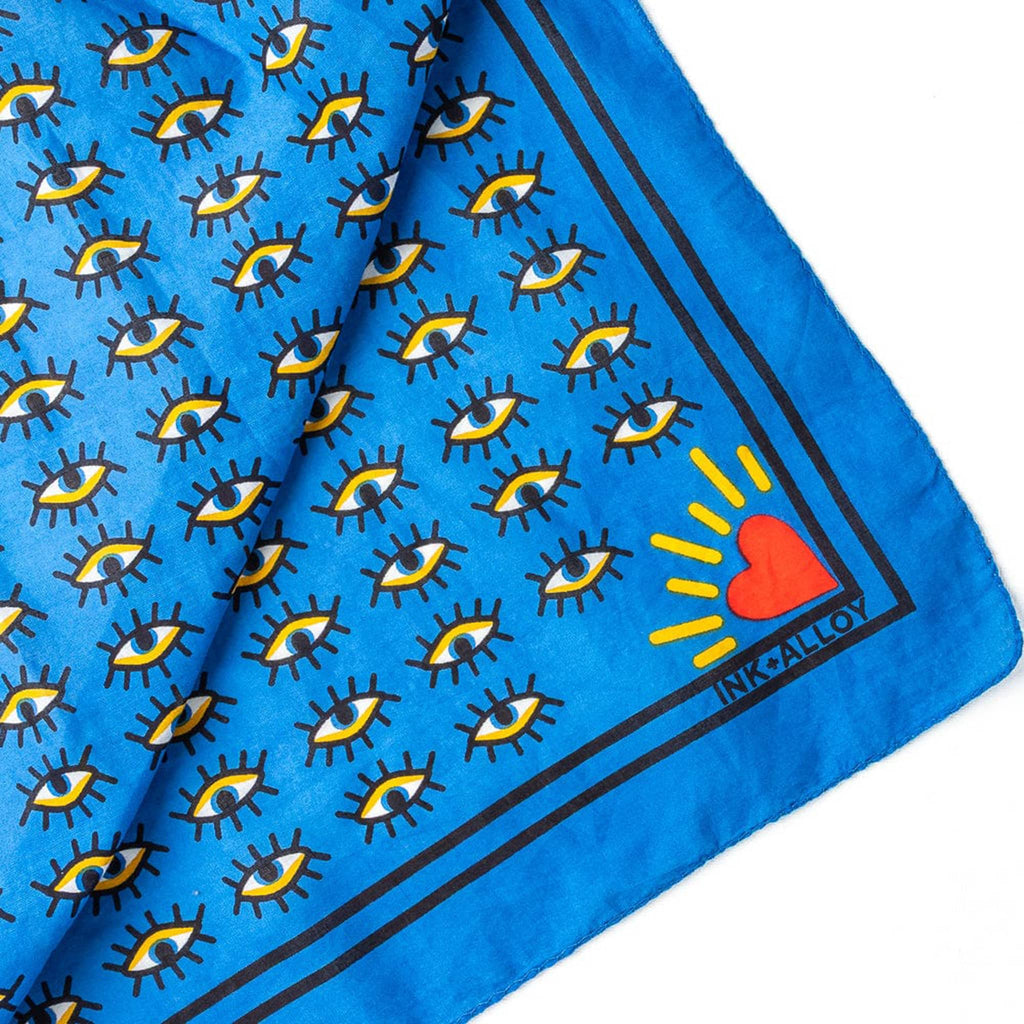 Corner of a blue scarf with eye pattern and red heart logo on a white background.