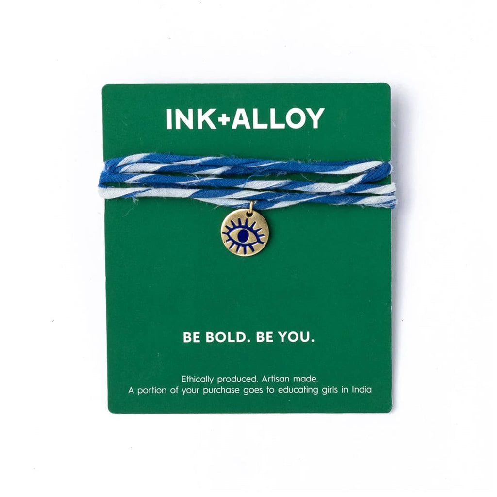 Bracelet with blue and white twisted cord and brass eye charm on a green card with 'INK+ALLOY' branding.