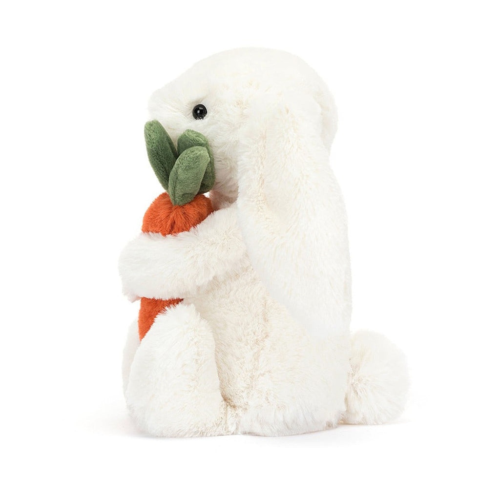 Jellycat Bashful Bunny plush toy in white, holding an orange plush carrot with green suedey top, side view.
