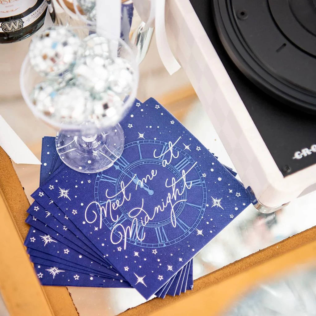 Jollity and Co midnight blue small paper party napkins with "meet me at midnight" in silver foil, a stack shown with glass filled with disco balls sitting on top.