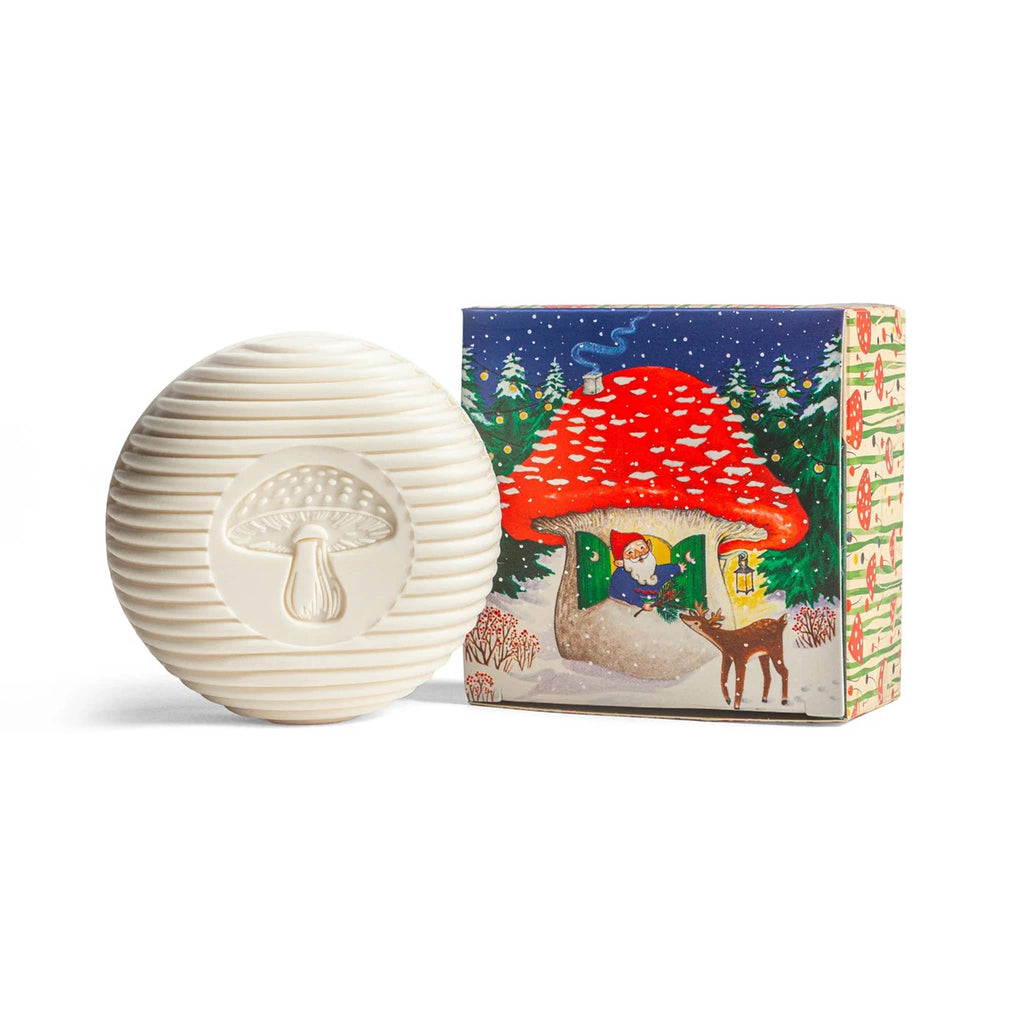 Round bar soap with mushroom imprint next to a box with a gnome in a mushroom greeting a deer in a snowy winter scene.