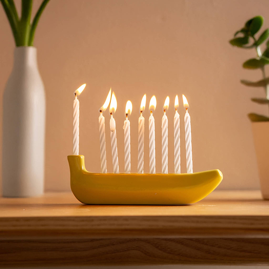Banana-shaped mini menorah with lit candles on a wooden surface, flanked by plants.