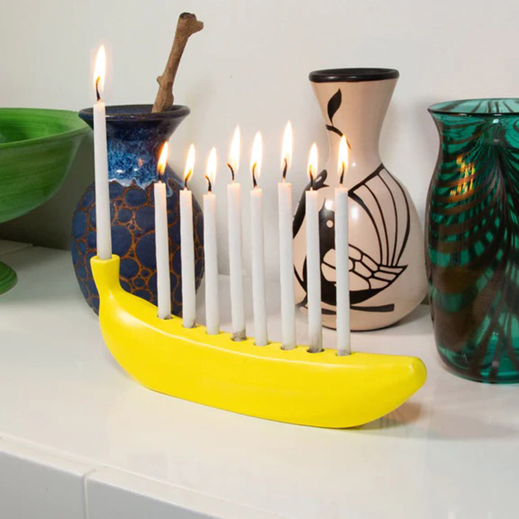 Kikkerland Banorah, yellow ceramic menorah in the shape of a banana, with lit candles with vases in the background.