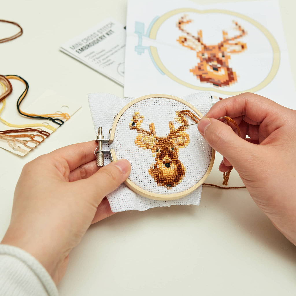 Person cross-stitching a deer design on white fabric with embroidery kit contents.