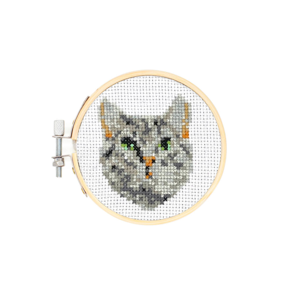 Cross-stitched gray cat head in an embroidery hoop on a white background.