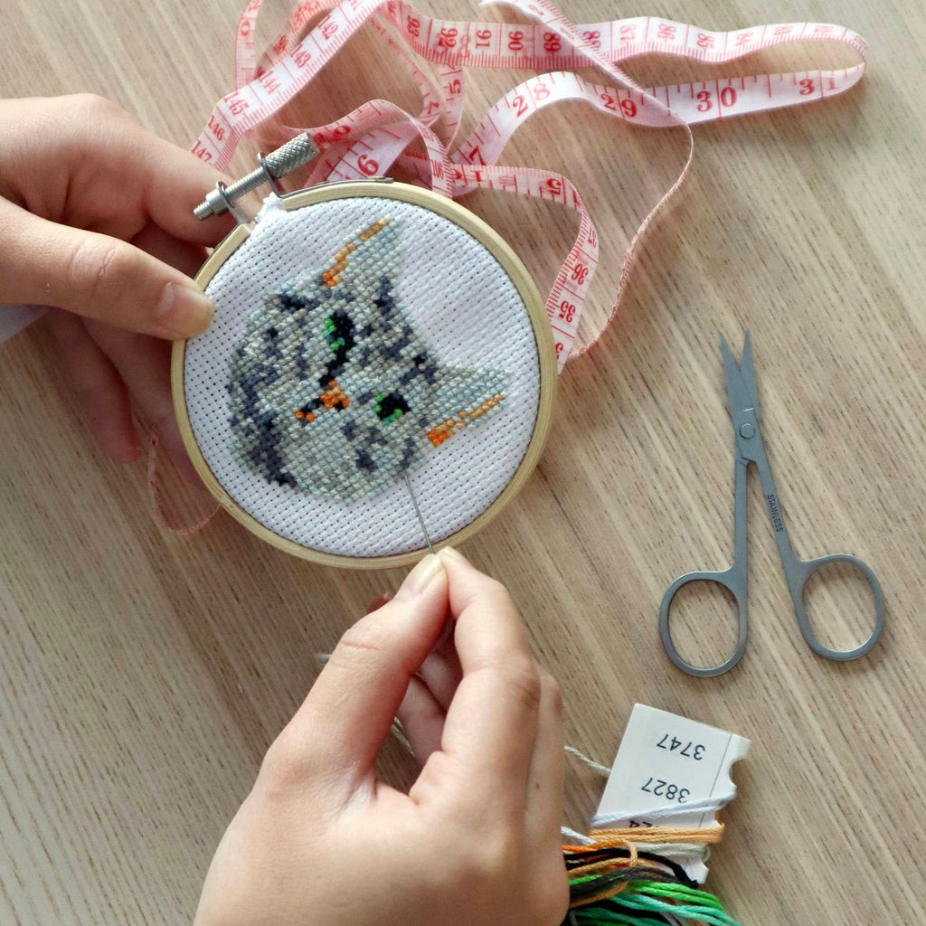 Person holding a cross-stitched hoop with a cat design on a wooden surface, with scissors and thread in the background.