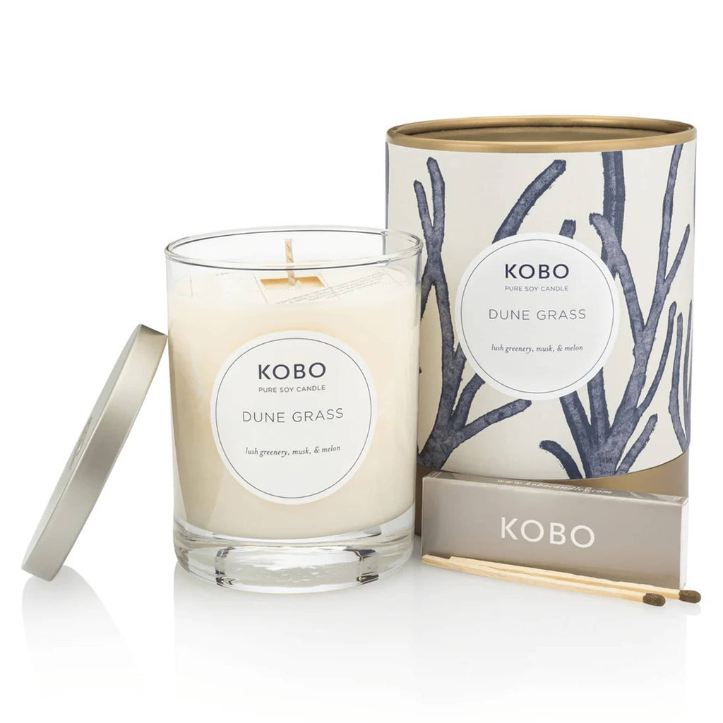 KOBO 11 ounce Dune Grass candle, part of the Coastal Collection, in clear glass vessel with metal lid next to blue and white illustrated canister packaging with a pack of wooden matches.