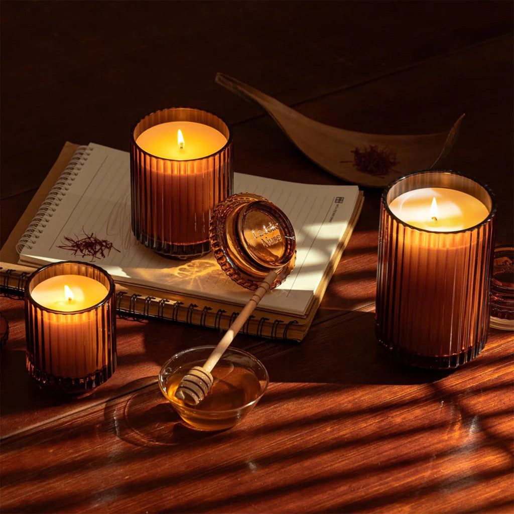 La Jolie Muse Amelie Saffron Oud scented soy wax candles in ribbed amber glass vessels with lids, in 3 sizes, wicks lit with a pad of paper and dish of honey..