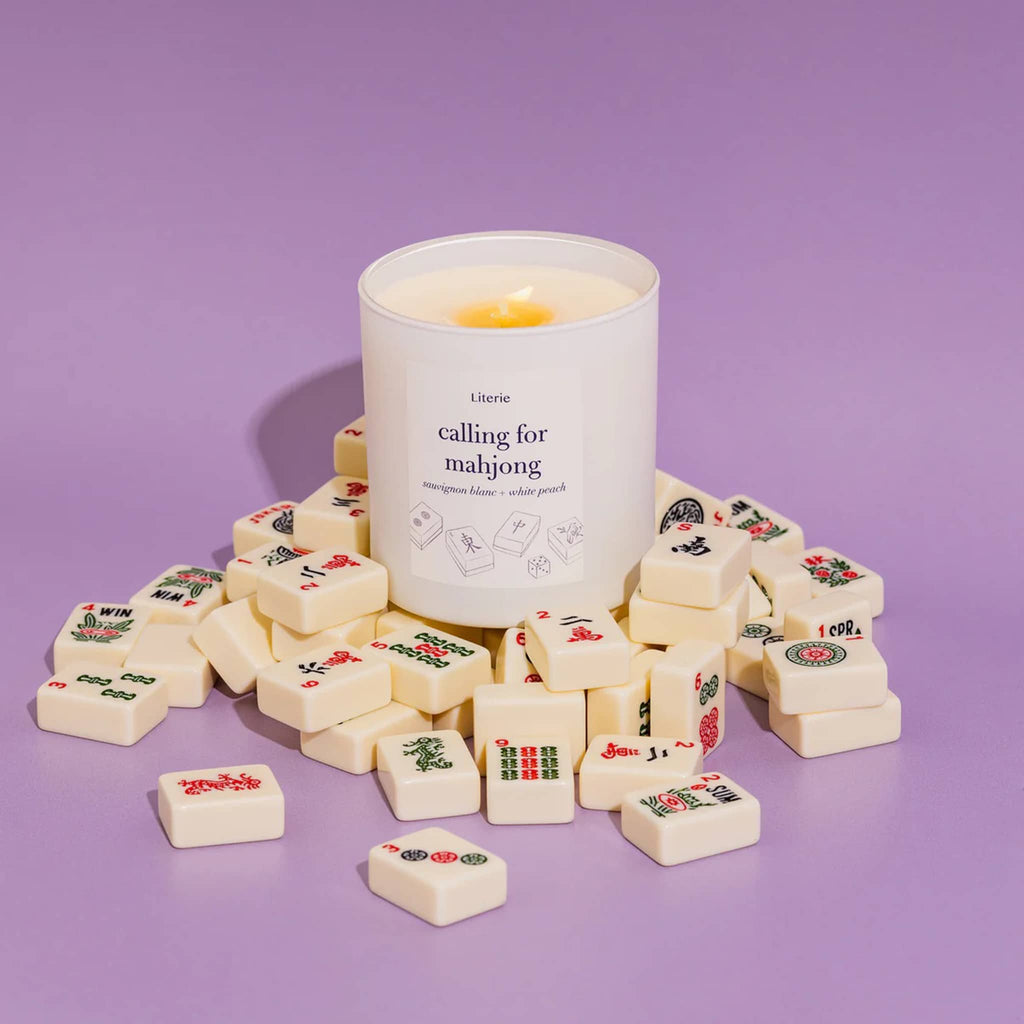 Literie Calling for Mahjong sauvignon blanc and peach scented candle in matte white vessel, wick lit, with mahjong tiles on a purple background.
