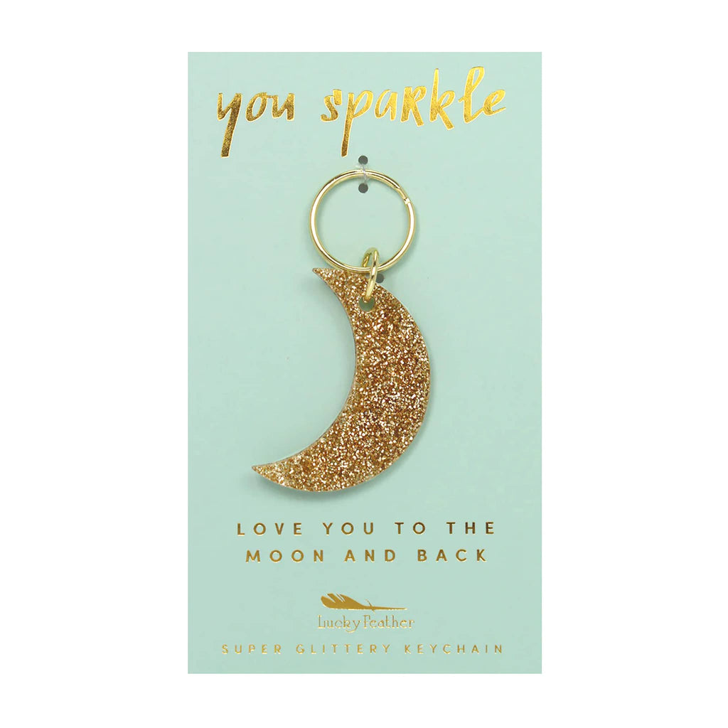 Gold glittery keychain shaped like a crescent moon on a light green card with text.