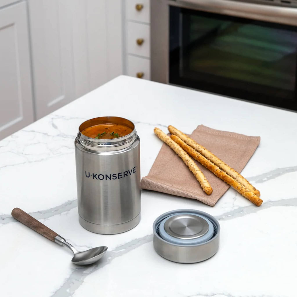 U-Konserve 18 ounce insulated food jar filled with soup, lid off with a spoon and breadsticks on a kitchen counter.