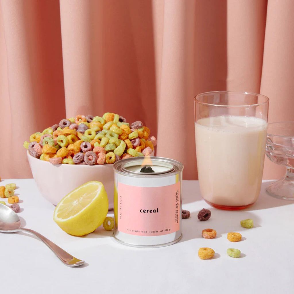 Mala the Brand Cereal Scented 8 ounce candle in metal tin with wood wick and a dusty pink label, wick is lit. Candle is sitting in front of a bowl of fruit flavored o shaped cereal in a light pink bowl and a glass of milk with a half lemon, a spoon and a dusty pink curtain behind.