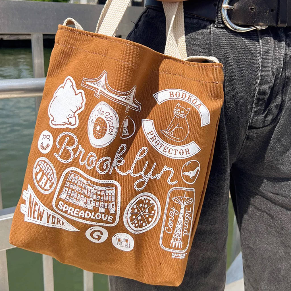 Brown tote bag with white Brooklyn graphics and text, with white webbed handles and shoulder strap, being carried by a person.