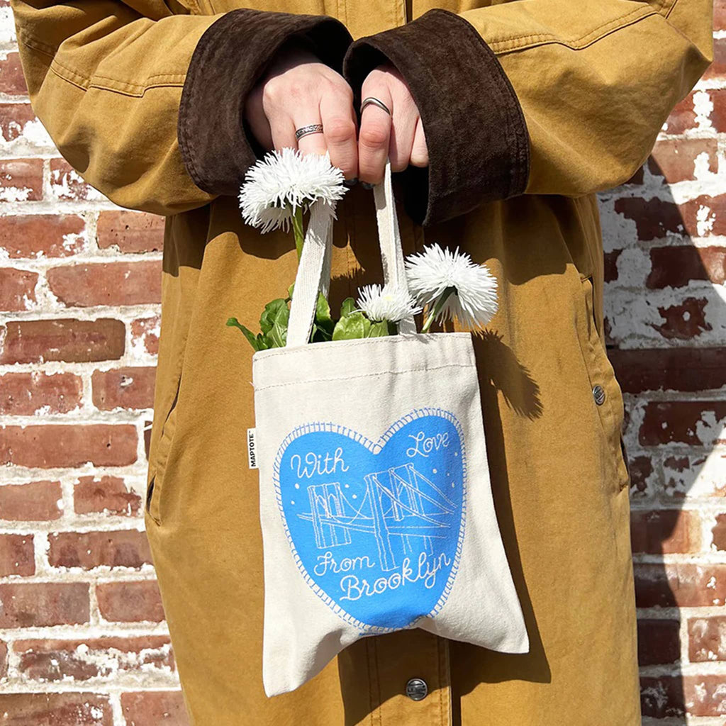Person holding a mini canvas tote bag with white flowers against a brick wall.