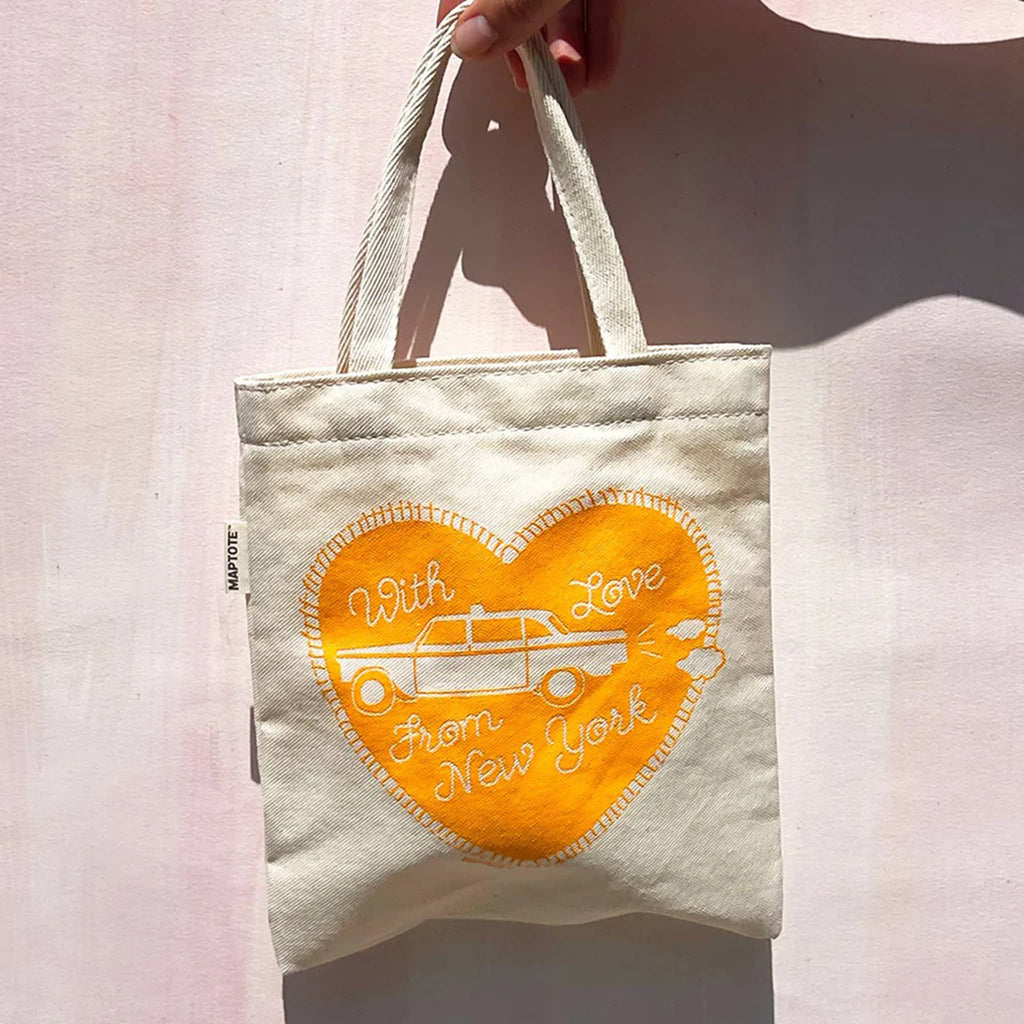 Small canvas tote bag with a yellow heart design with taxi and "with love from new york" being held by a hand against a pink wall background.