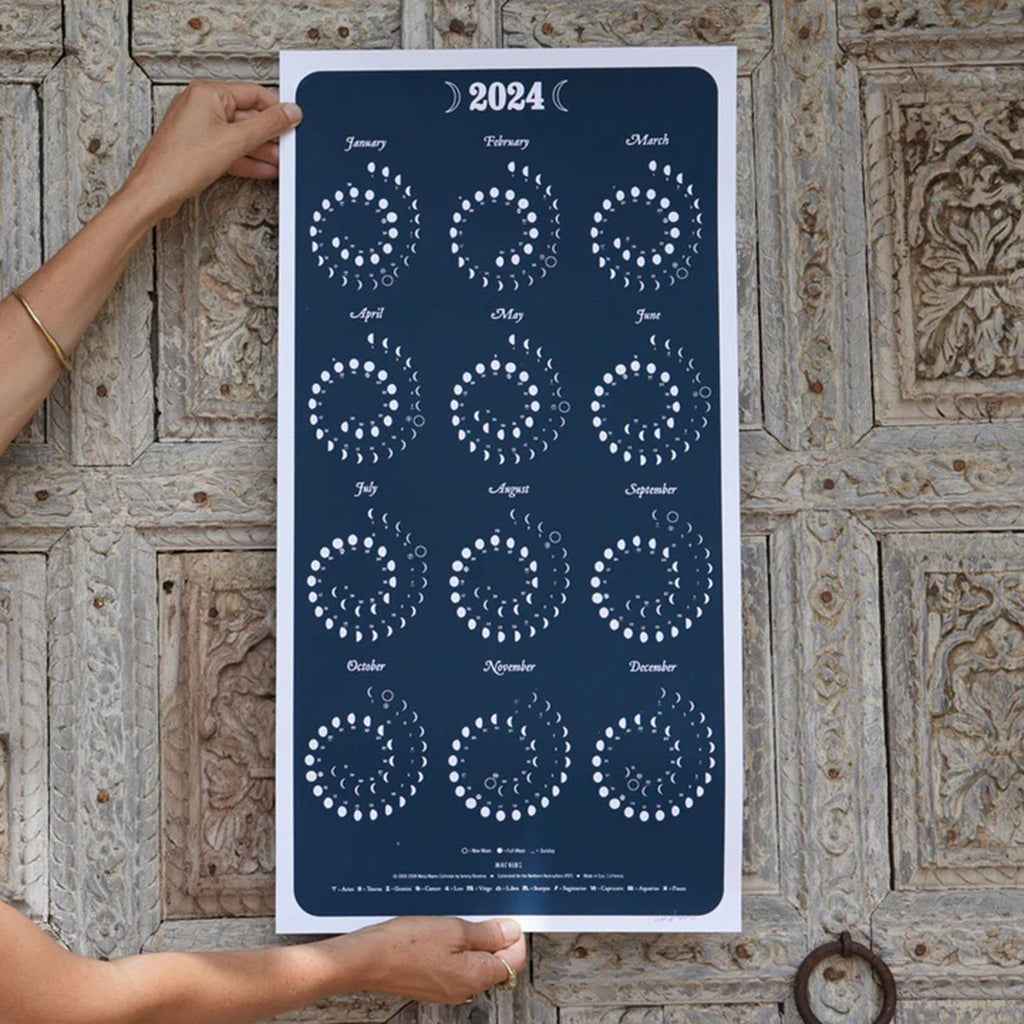 Large vertical rectangular poster showing 2024 phases of the moon by month in white on a navy blue background. Poster is held up by hands against a blue tinted textured wood surface.