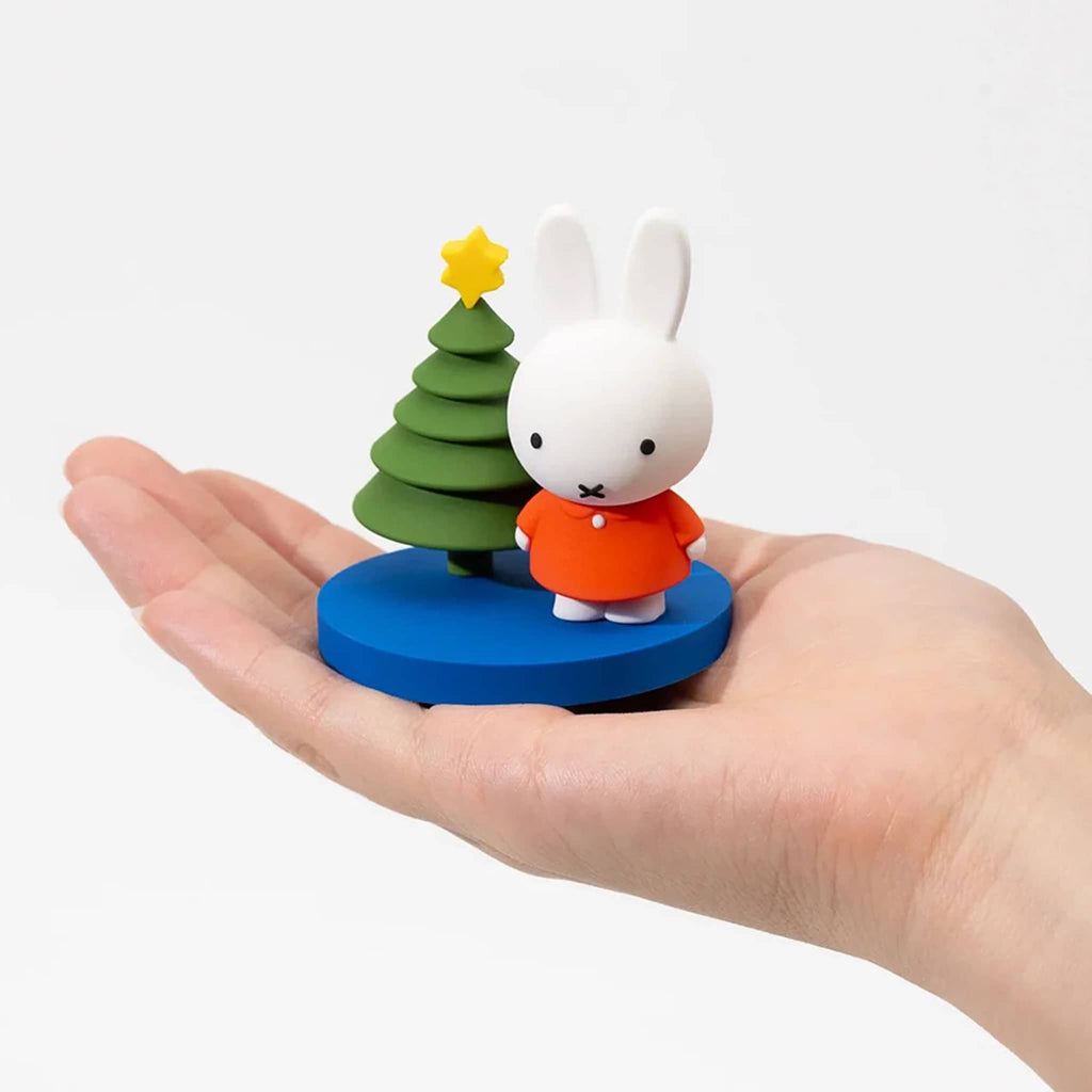 Small figurine of a white rabbit in an orange coat standing next to a green tree with a yellow star on a blue base, held in a hand against a white background.