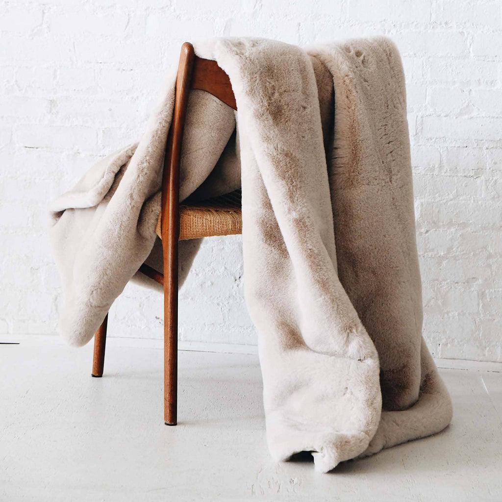 MERSEA Cozy Cabin Faux Fur Throw Blanket in oatmeal cream on a chair.