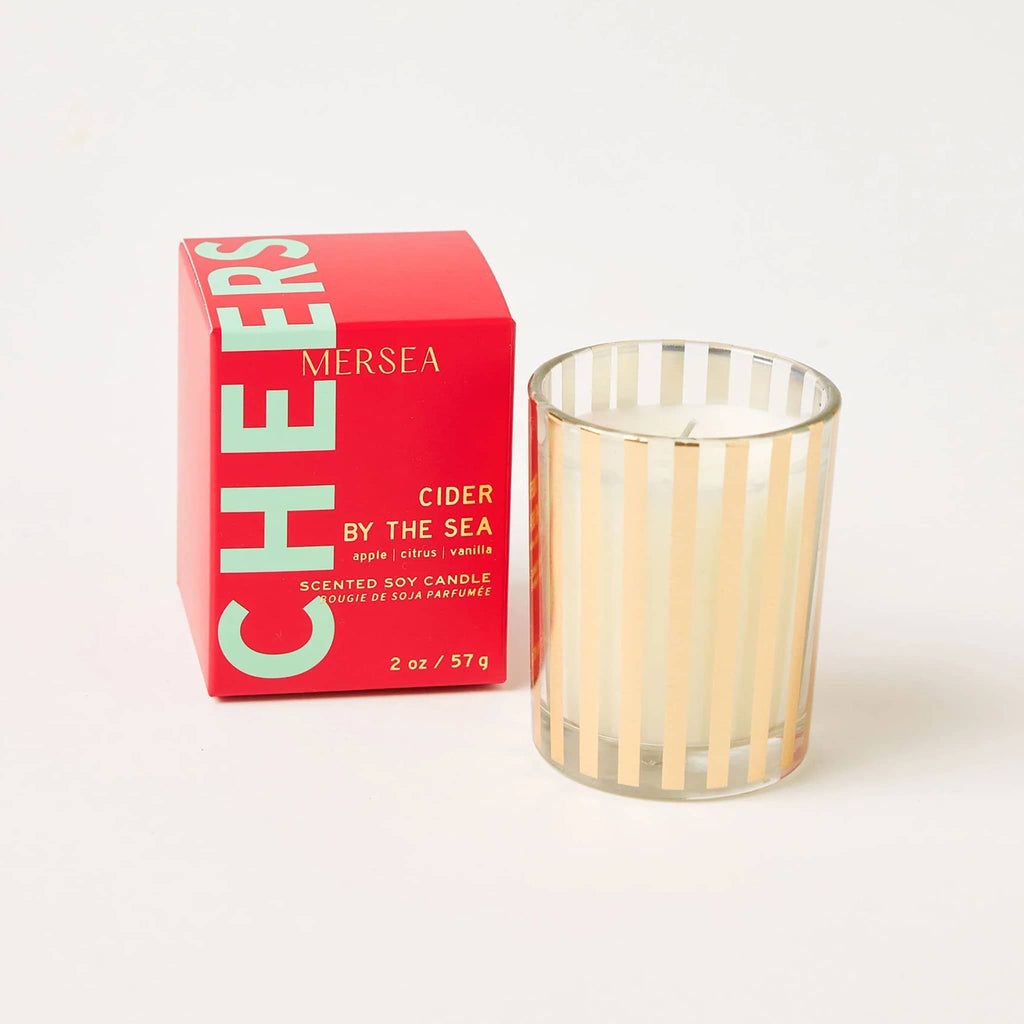 Mer-Sea Good Tidings Cider by the Sea scented votive candle in gold striped vessel, beside red box packaging.