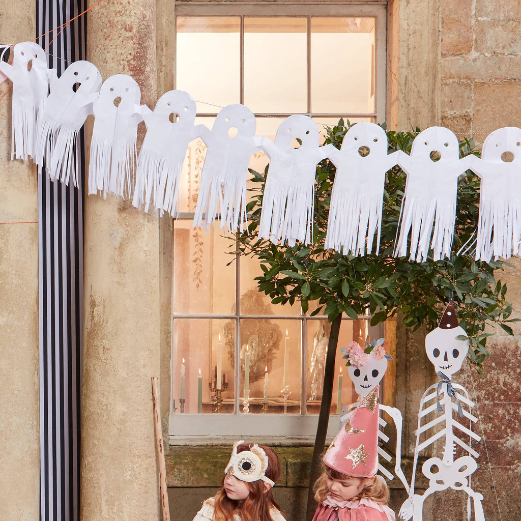 Decorative ghost banner with tassels hanging above a window with candlelight, children in costumes below.