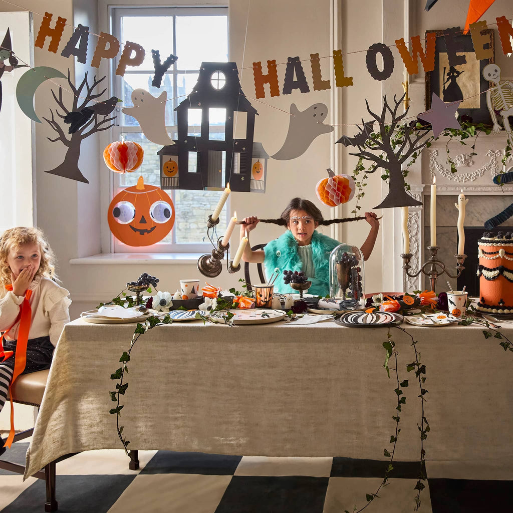 Children at a Halloween-themed table with decorations and a 'Happy Halloween' banner with Halloween icons such as ghosts and pumpkins.
