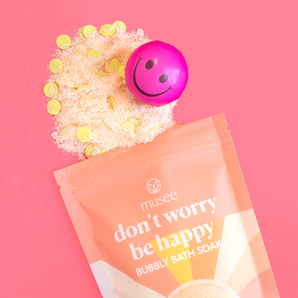 Musee Bath Don't Worry Be Happy bubbly bath soak in yellow and orange resealable bag with sun illustration and smiley face stress ball surprise on a pink background.