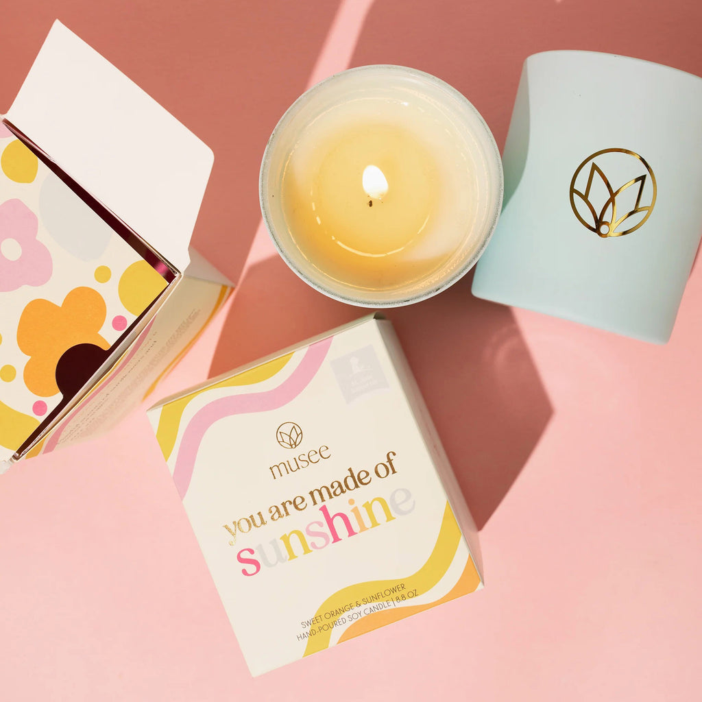 Musee x St. Jude's Children's Hospital You Are Made of Sunshine sweet orange and sunflower scented soy wax candle in matte sky blue glass vessel with musee logo in gold foil, with box packaging and a lit candle, overhead view on salmon pink background.
