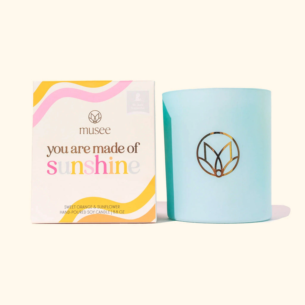 Musee x St. Jude's Children's Hospital You Are Made of Sunshine sweet orange and sunflower scented soy wax candle in matte sky blue glass vessel with musee logo in gold foil, beside the colorful illustrated box packaging.